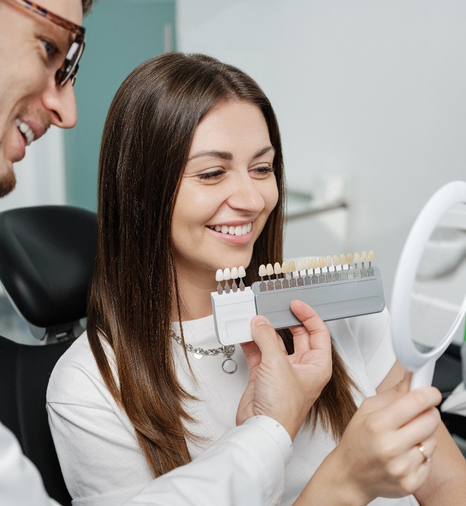 a woman looking into a mirror while a dentist holds a shade chart up to her smile