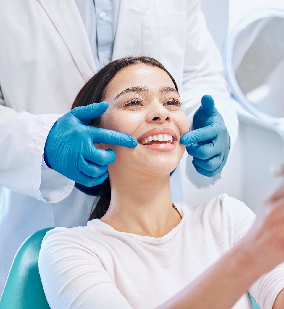 Dentist touching woman’s cheeks as she smiles at her reflection in handheld mirror